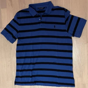 Blå randig Polo Golf piké XL - Snygg blå piké från Polo Golf Ralph Lauren i XL med svarta horisontella ränder och klassisk pikékrage. Pikén har korta ärmar, två knappar vid halsen och den ikoniska Polo-loggan broderad på bröstet. Perfekt för dig som gillar sportig och stilren look.