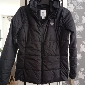 Svart Adidas pufferjacka med huva - Svart pufferjacka från Adidas med huva och klassiska vita ränder längs sidorna. Jackan har dragkedja framtill, quiltad design och Adidas logga på bröstet. Tillverkad i Primegreen-material och passar perfekt när du vill hålla dig varm och sportig.