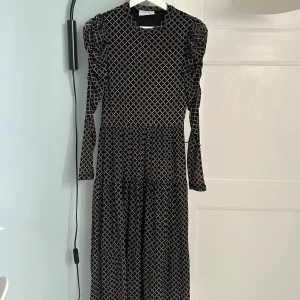 Svart långklänning med mönster från Neo Noir - Snygg långklänning från Neo Noir i svart med beige rutigt mönster med guldiga glitter detaljer. Klänningen har puffade axlar, lång ärm i elastisk mesh och rund halsringning. 