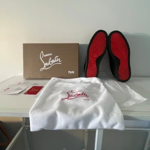 Christian Louboutin svarta sneakers med nitar - Svarta sneakers från Christian Louboutin i mocka med ikoniska röda sulor och svarta nitar över tån. Skorna har snörning och en rund tå, vilket ger en edgy och lyxig vibe. Perfekta för dig som vill sticka ut med exklusiv streetstyle. Storlek 40 allt finns. Box,papper,kvitto och kort.