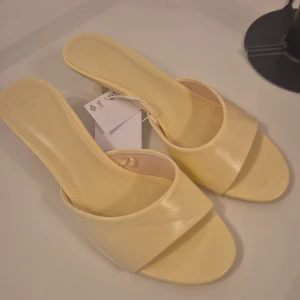 Gula sandaler med klack från Bershka - Snygga ljusgula sandaler från Bershka med låg klack och öppen tå. Skorna har en bred rem över foten och är tillverkade i syntetmaterial med en stilren, minimalistisk design. Perfekta för att lyfta din look med en pop av färg.