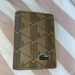 Beige plånbok från Lacoste med mönster - Snygg beige plånbok från Lacoste med geometriskt mönster och klassiska krokodilloggan framtill. Plånboken är i skinn och har flera fack för kort på insidan. Perfekt för dig som vill ha en stilren och unik accessoar.