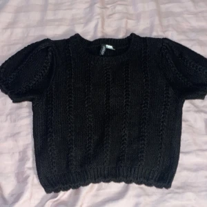 Svart stickad tröja från H&M Divided - Svart stickad tröja från H&M Divided i storlek S. Tröjan har korta puffärmar och ett snyggt hålmönster längs hela plagget. Ribbad kant nedtill och vid ärmslut. Perfekt att styla med jeans eller kjol för en trendig look.