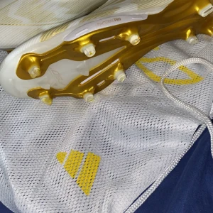 Adidas F50+ fotbollsskor vit/guld - Säljer ett par Adidas F50+ fotbollsskor i vitt med snygga gulddetaljer och klassiska tre ränder på sidan. Skorna har en lätt ovandel i syntetmaterial och guldiga dobbar för grymt grepp på planen. Perfekta för dig som vill sticka ut med stil på matchen. Lite sumutsiga bara har kört en 1 match