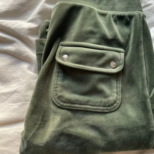 Olivgröna mjukisbyxor Juicy Couture - Olivgröna mjukisbyxor från Juicy Couture i mjuk velour med bred resår och snörning i midjan. Byxorna har två fickor med lock och silverfärgade knappar bak samt broderad JC-logga fram. Perfekta för chill eller streetwear. Snörena i midjan har som små silver detaljer, och en utav dem lossnar ibland!! Ska absolut försöka laga innan jag postar!!❣️❣️