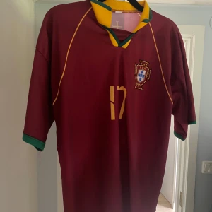Portugal C. Ronaldo 17 fotbollströja - Säljer en klassisk Portugal fotbollströja med C. Ronaldo och nummer 17 på ryggen. Tröjan är vinröd med gula detaljer, gröna muddar och gul/grön krage. Officiellt landslagsmärke på bröstet. Perfekt för dig som älskar fotboll och Ronaldo. Tyvärr är den för liten för mig och det är därför jag säljer den.