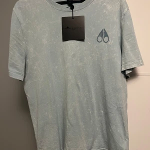 Ljusblå Moose Knuckles t-shirt - Snygg ljusblå t-shirt från Moose Knuckles med coolt tvättat mönster. Stor logga på ryggen och mindre logga framtill. Klassisk passform med korta ärmar och rund hals. Tillverkad i mjuk bomull som känns skön mot huden. Perfekt för en avslappnad och trendig look.