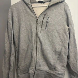 Grå hoodie från Sail Racing - Säljer en grå hoodie från Sail Racing med dragkedja framtill och logga på bröstet. Tröjan har huva, lång ärm och en extra bröstficka med dragkedja. Perfekt för dig som gillar sportig och avslappnad stil.
