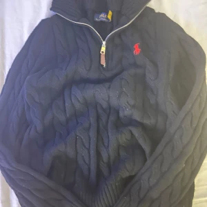 Svart kabelstickad tröja Polo Ralph Lauren - Kabelstickad mörkblå tröja från Polo Ralph Lauren med halv dragkedja och röd broderad logga på bröstet. Tröjan har hög krage och är tillverkad i mjuk ull, perfekt för kyliga dagar. Klassisk och stilren design som funkar till det mesta.