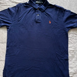 Marinblå Polo t-shirt från Ralph Lauren - Snygg marinblå piké från Polo Ralph Lauren i bomull. Klassisk krage med två knappar och orange broderad logga på bröstet. Perfekt för dig som gillar stilrena och tidlösa plagg. Passar dig som vill ha en clean look med lite preppy vibe.
