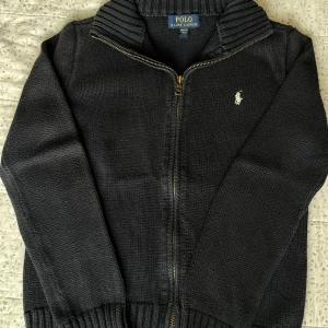 Svart stickad kofta från Polo Ralph Lauren - Mysig svart stickad kofta från Polo Ralph Lauren med dragkedja framtill och ribbad krage. Klassisk broderad logga på bröstet och ribbade muddar vid ärmslut och nederkant. Perfekt för lager-på-lager under kyliga dagar.