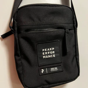 Peak Performance Axelremsväskan - Snygg svart axelväska från Peak Performance med justerbar rem och dragkedja framtill. Väskan har en stilren design med logga på framsidan och extra fack både fram och bak. Perfekt storlek för att bära med sig det viktigaste.