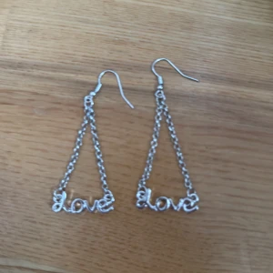 Silvriga örhängen med love-text - Snygga silvriga örhängen med kedjor som bildar en triangel och texten 'love' längst ner. Perfekta för att ge din look lite extra attityd och kärlek. Hängande modell som syns och sticker ut.