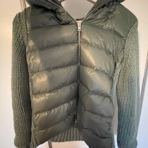 Grön pufferjacka/ cardigan från Herno med stickade ärmar - Snygg grön pufferjacka från Herno med stickade ärmar och huva. Jackan har quiltad framsida i glansigt material och dragkedja hela vägen. Perfekt för kyliga dagar när du vill vara både varm och stilren. Skriv till mig för fler bilder! Jackan är i perfekt nyskick och använd bara några gånger. 