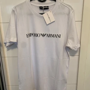 Vit t-shirt från Emporio Armani - Snygg vit t-shirt från Emporio Armani med svart logga tryckt på bröstet. Klassisk rund halsringning och korta ärmar. Tillverkad i mjuk bomull med en liten del lycra för extra stretch. Perfekt för dig som gillar stilrena och exklusiva plagg.