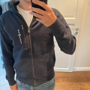 Mörkblå hoodie från Sail Racing - Säljer en mörkblå hoodie från Sail Racing med dragkedja framtill och en extra bröstficka med zip. Tröjan har huva, långa ärmar och en broderad logga på huvan. Väldigt gosig och varm perfekt nu när börjar bli lite kallare! Nypris 1500