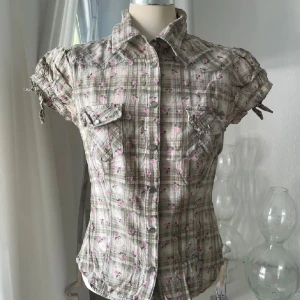 Kortärmad rutig skjorta med blommor - Kortärmad skjorta i beige och grått med rutigt mönster och små rosa blommor. Skjortan har knappar framtill, två bröstfickor med lock och tryckknappar samt knytband på ärmarna. Figurnära passform och klassisk krage. Perfekt för dig som gillar en söt och avslappnad stil.