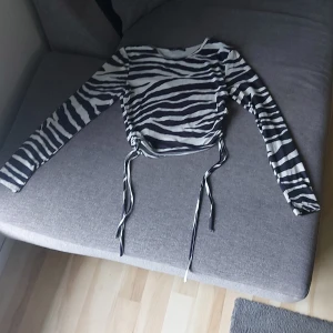 Zebramönstrad långärmad topp - Snygg långärmad topp med zebramönster i svart och vitt. Toppen har trendiga snörningsdetaljer på sidorna som gör den extra cool och croppad. Perfekt för dig som vill sticka ut med en djurinspirerad look.