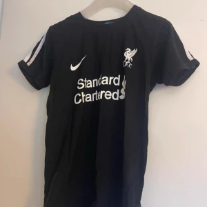 Svart Liverpool fotbollströja Itali - Svart fotbollströja med Liverpool FC-logga och Nike-swoosh på bröstet, samt vit Standard Chartered-tryck. Tröjan har vita ränder längs axlarna och korta ärmar. Materialet är lätt och andas, perfekt för träning eller match. Rund hals och klassisk passform.