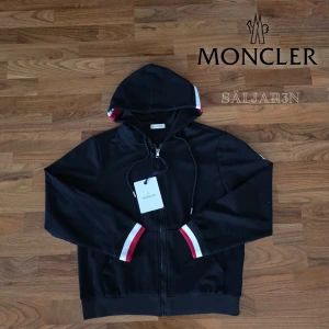 Moncler Hoodie - Exklusiv Moncler hoodie i svart, storlek S. Nyskick med taggar! Elegant design med hel dragkedja och klassiska Moncler-detaljer i rött, vitt och blått på muddar och huva. Ikoniskt logomärke på ärmen för en exklusiv touch. Huva med dragsko och fickor för en modern, funktionell stil. Tillverkad i mjukt och slitstarkt tyg som ger både komfort och premiumkänsla. En tidlös lyxig hoodie som lyfter varje outfit. 📦 Snabb leverans – skickas samma dag!