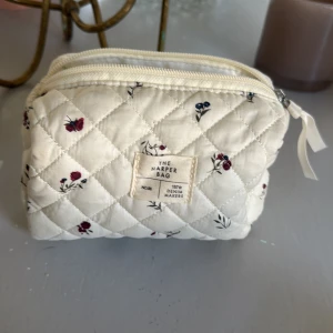 Quiltad blommig necessär från The Harper Bag - Superfin quiltad necessär i ljusbeige bomull med små blommor i rött och blått. Väskan har dragkedja upptill och en mjuk, vadderad känsla. Perfekt för smink eller småprylar. Söt detalj med etikett framtill.