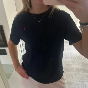 Mörkblå t-shirt från Ralph Lauren - Snygg mörkblå t-shirt från Ralph Lauren med klassisk röd logga broderad på bröstet. Modellen är normal i passformen och har korta ärmar. Materialet är mjuk bomull som känns skönt mot huden. Perfekt för dig som gillar stilrena och enkla plagg.