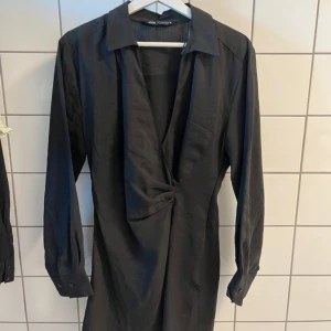Svart omlottklänning från Zara - Snygg svart kortklänning från Zara med omlottdetalj framtill och v-ringning. Klänningen har långa ärmar med manschett och krage, samt smock i ryggen. Passar S, använd 1 gång