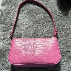 Rosa axelväska i croco-imitation - Säljer en snygg rosa axelväska med croco-mönstrad yta och silvrig dragkedja. Väskan har en stilren form och ett rymligt svart foder inuti. Perfekt för dig som vill sticka ut med en färgglad accessoar.
