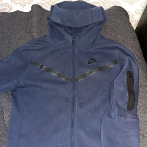 Marinblå Nike hoodie XL - Säljer en marinblå hoodie från Nike i storlek XL (158-170 cm). Tröjan har hel dragkedja, huva och svarta detaljer på bröstet och ärmen. Perfekt för chill eller träning, med långärmad passform och mjukt material som känns skönt mot huden. Skicket är bra och anledningen till att jag vill sälja den är att den är för liten för mig. 