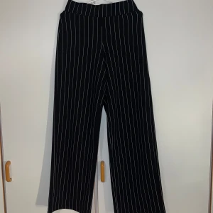 Svarta randiga byxor från Gina Tricot - Snygga svarta byxor med vita vertikala ränder från Gina Tricot. Byxorna har en rak och loose passform med hög midja och vida ben. Tillverkade i ett mjukt material med stretch som gör dem riktigt bekväma. Perfekta för en trendig och chill look.