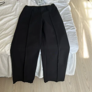 Svarta kostymbyxor från Gina  - Snygga svarta kostymbyxor med raka ben och pressveck. Byxorna har hällor för bälte och en klassisk, stilren look som passar till många olika outfits. Perfekta för dig som gillar en clean och dressad stil.