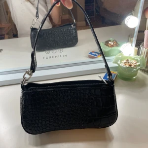 Svart axelväska i croco-imitation - Snygg svart axelväska med croco-mönstrad yta och silvriga metalldetaljer. Väskan har en smidig dragkedja upptill och ett stilrent, avlångt format som passar perfekt till dina essentials. Enkel att matcha med olika outfits och ger en trendig touch.