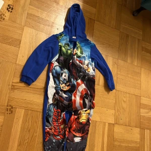 Marvel Avengers blå hoodie onepiece - Blå onepiece med huva och långa ärmar, prydd med stort Avengers-tryck framtill med Hulk, Thor, Captain America och Iron Man. Mjuk och skön, perfekt för myskvällar. Dragkedja framtill och ribbade muddar vid ärmslut.