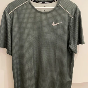 Grå Nike Dri-Fit t-shirt - Nike Dri-Fit t-shirt i grått med diskret rutigt mönster och reflekterande detaljer vid axlarna. Kortärmad och tillverkad i lätt, ventilerande polyester som håller dig torr under löpningen. Swoosh-logga på bröstet och rund halsringning.