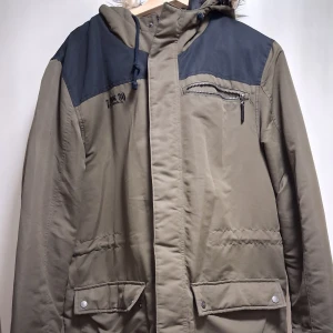 Parkajacka från Z.ink med fuskpäls - Parkajacka från Z.ink i oliv  och mörkgrått med huva och fuskpäls. Jackan har dragkedja, tryckknappar, flera fickor och quiltade detaljer. Perfekt för kalla dagar med sin långa modell och varma känsla.