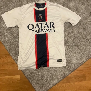 PSG Mbappé 7 vit matchtröja Nike - Paris Saint-Germain fotbollströja med Mbappé och nummer 7 på ryggen. Vit med bred marinblå rand och röda detaljer på framsidan, PSG-emblem och Nike-logga. Qatar Airways tryck. Tillverkad i Dri-FIT material som andas, perfekt för träning eller match.