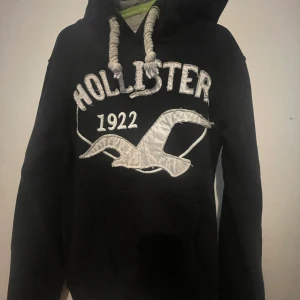 Svart hoodie från Hollister med tryck - Svart hoodie från Hollister med stor vit broderad logga och texten 'Hollister 1922' på bröstet. Tröjan har grova snören i huvan, känguruficka och är i mjukt material. Perfekt för dig som gillar klassisk college-stil. Obs vet inte om det är äkta hollister. Den är i storlek m men väldigt liten i storlek så passar xs-xxs. Den är lite miss färgad som man ser på bilden.