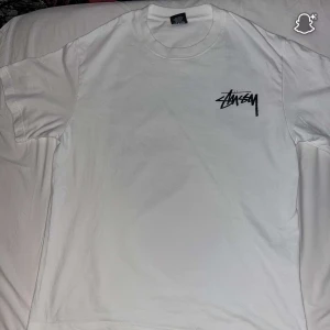 Vit Stüssy t-shirt med tryck - Vit t-shirt från Stüssy i bomull med svart logga på bröstet och stort tryck på ryggen med rosa tärningar och Stüssy-text. Klassisk rund hals och korta ärmar, perfekt för en streetwear-look.