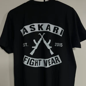 Svart Askari Fight Wear t-shirt - Svart t-shirt från Askari Fight Wear med stor vit logga och tryck av korsade gevär på både bröstet och ryggen. Klassisk rund hals och korta ärmar. Perfekt för dig som gillar streetwear och vill sticka ut med en bold design.