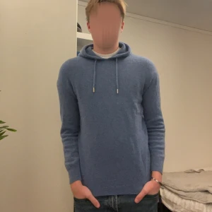 Blå hoodie i kashmir med huva - Mysig blå hoodie i kashmir med huva och dragsko. Tröjan har lång ärm och ribbade muddar vid ärmslut och nederkant. Perfekt för dig som gillar en stilren och enkel look med premiumkänsla. Storlek S