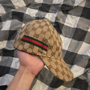Beige  keps med GG-mönster - Säljer en beige keps med klassiskt GG-monogram över hela kepsen och en grön-röd rand på sidan. Justerbar metallspänne baktill. Snygg och lyxig accessoar som lyfter din stil. Skriv innan ni köper