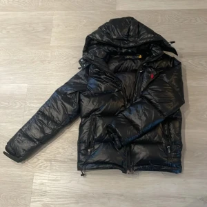 Svart pufferjacka med huva(hjälper en vän) - Snygg svart pufferjacka från Polo Ralph Lauren med glansig finish och huva. Jackan har dragkedjor på fickorna och en liten röd logga på bröstet. Perfekt för kalla dagar och riktigt skön att bära.