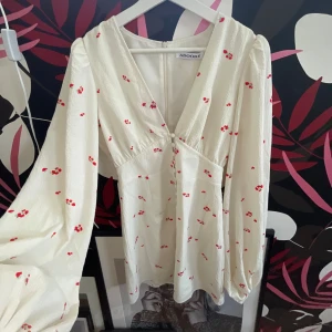 Vit blommig kortklänning från Adoore - Supersöt vit kortklänning från Adoore med små röda blommor, v-ringning och långa puffärmar. Klänningen har fina klädda knappar framtill och är gjord i ett lätt, strukturerat tyg som känns luftigt och fräscht. Perfekt för varma dagar och somriga vibes!