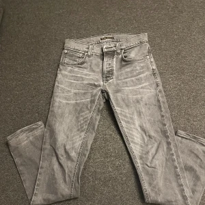 Grå jeans från Nudie Jeans, 29/32 - Snygga grå jeans från Nudie Jeans med klassisk femficksdesign och svarta broderade detaljer på bakfickorna. Jeansen har raka ben och normal passform, tillverkade i mjukt bomullstyg. Perfekta för en avslappnad och stilren look. (Model: Grim Tim, storlek: W29 L32). Vid eventuella frågor skriv gärna!