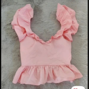 Rosa volangtopp med knytband - Supersöt rosa topp med volanger på axlarna och nertill. Djup v-ringning och knytband i ryggen. Perfekt för sommaren! Kommer med prislappen kvar, alltså aldrig använd! 