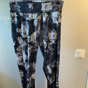 Mönstrade träningstights från Energetics - Säljer ett par snygga träningstights från Energetics i storlek small. Tightsen har ett svart, grått och vitt blommigt mönster och är tillverkade i ett stretchigt syntetmaterial med hög midja. Perfekta för gymmet eller löprundan!