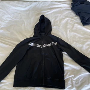 Svart kofta från Nike med dragkedja - Svart kofta från Nike med hel dragkedja och vit logga/mönster över bröstet. Kofta har huva och två fickor framtill. Perfekt för en sportig och avslappnad stil.