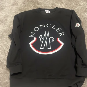 Svart sweatshirt från Moncler - Säljer en svart sweatshirt från Moncler med stort tryck på bröstet och logga på ärmen. Tröjan har rund halsringning och ribbade muddar vid ärmslut och nederkant. Perfekt för en stilren och sportig look.