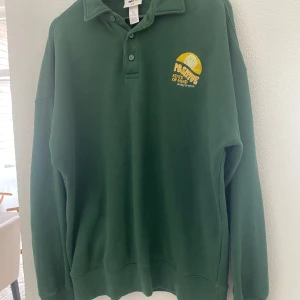 Grön pikétröja från H&M x Smiley Originals - Grön långärmad pikétröja från H&M x Smiley Originals med broderad sol och texten 'Positive State of Mind' på bröstet. Klassisk krage och knäppning framtill. Perfekt för en avslappnad stil.