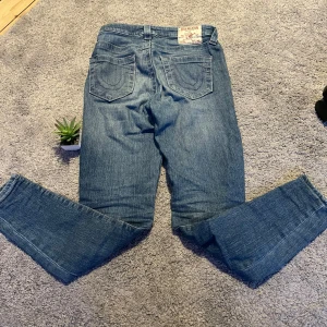 Blå jeansbyxor från true religion - Säljer ett par trendiga blå jeans från true religion med fem fickor och raka ben. Byxorna har normal passform. Perfekta för dig som gillar stockholmstil,grisch stil och vill ha ett par jeans som är sköna,stiliga 
